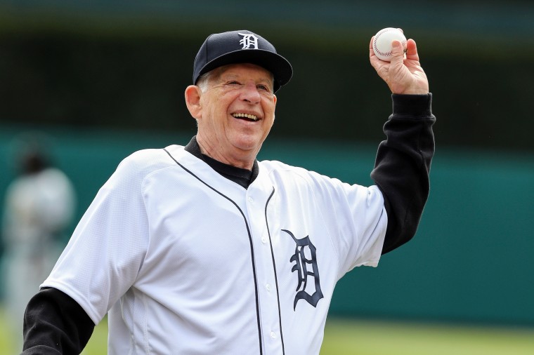An elderly Mickey Lolich smiles as he throws a baseball with his left hand. He's wearing a white and navy blue Detroit Tigers uniform.