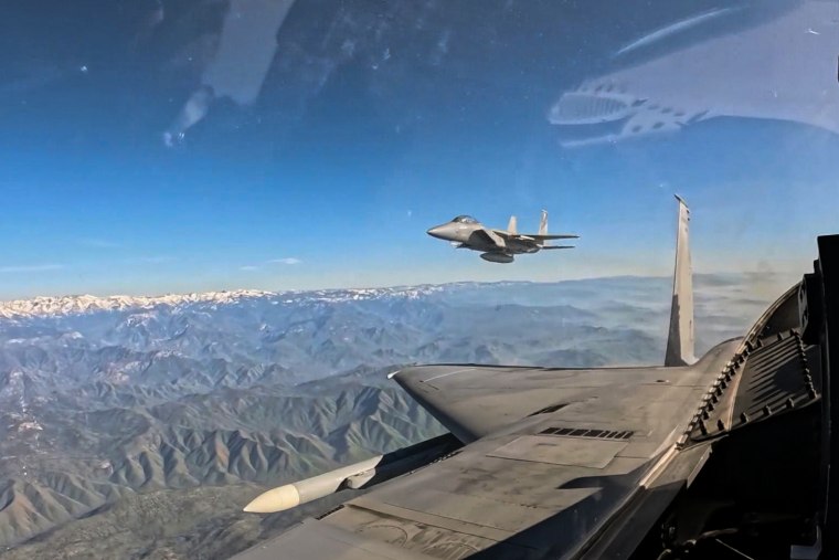 Two fighter jets fly high above a craggy mountain range with snow-capped peaks.