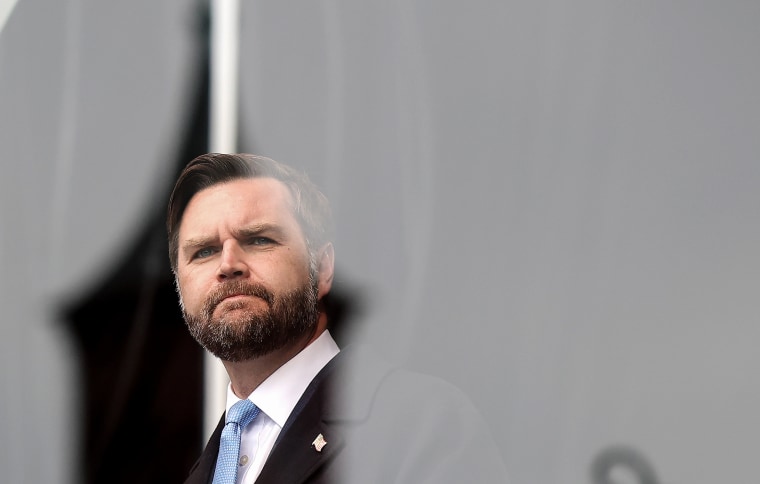 JD Vance appears behind a glass partition while onstage at an outdoor rally.