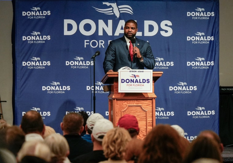 Byron Donalds holds a microphone while standing at a wooden lectern. A backdrop behind him reads DONALDS FOR FLORIDA.