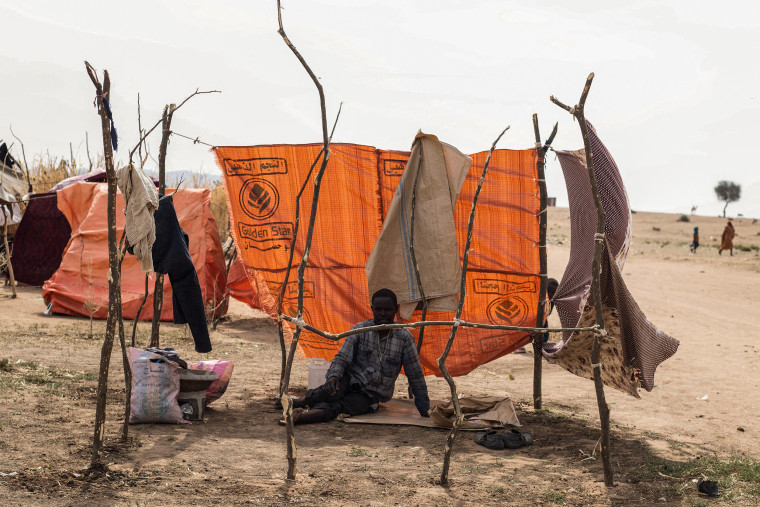 A young man sits on a blanket on the bare ground surrounded by tarps and blankets.