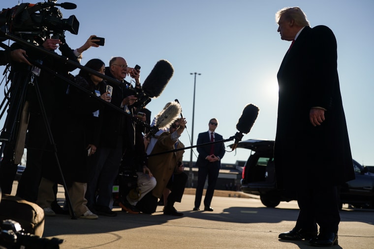 Donald Trump stands opposite a large group of reporters on a tarmac. They are silhouetted by the sunlight behind his head.