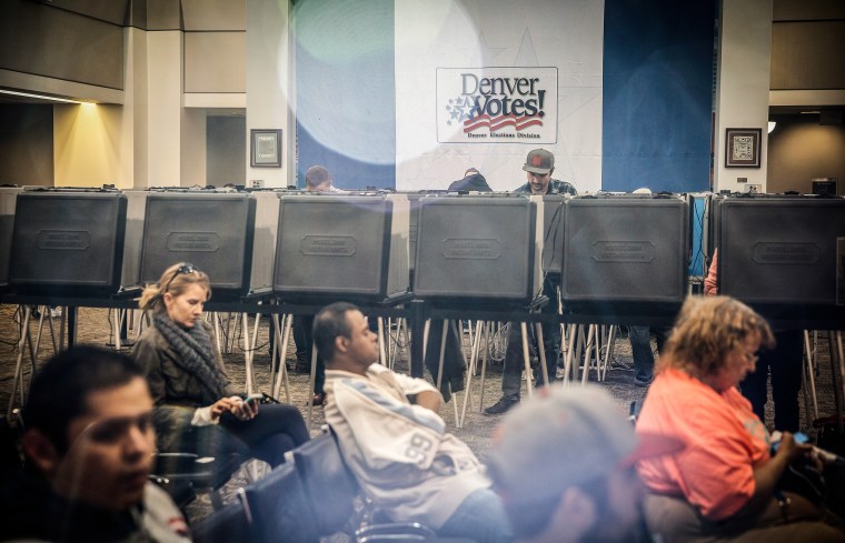 Several people stand at black plastic voting booths behind another group of people seated in black chairs. A sign on the wall reads DENVER VOTES!
