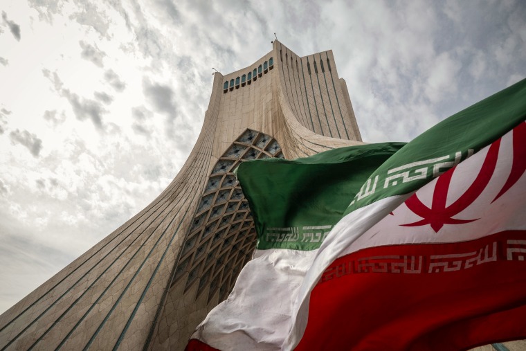 The green, white and red national flag of Iran flutters in front of a looming concrete tower.