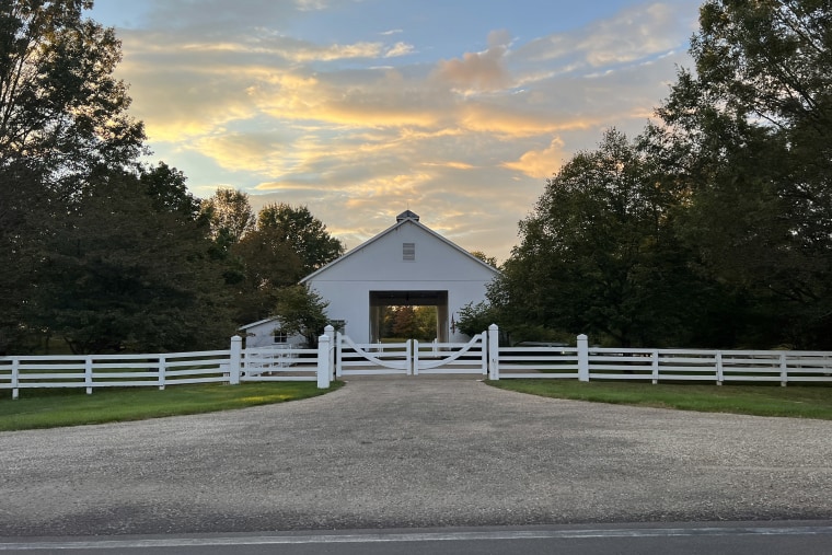 A white barn structure with a drive-thru opening on each side sits behind a white picket fence at sunset.