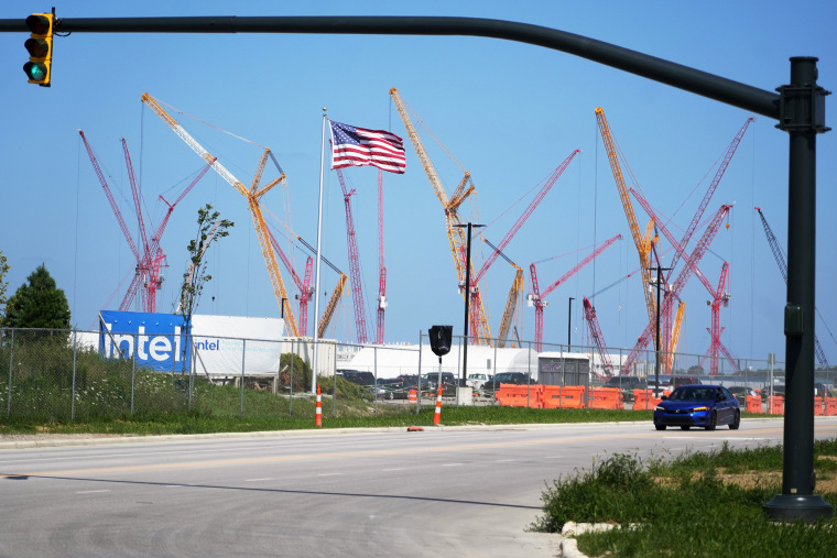 An American flag flutters in front of a large group of construction cranes, their arms extended toward the sky.