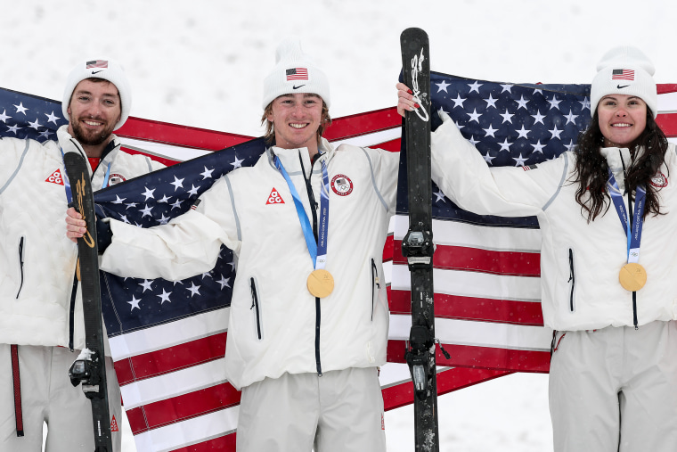 Three Olympic skiers stand together with gold medals around their necks. Two of them are holding a ski in their right hands.