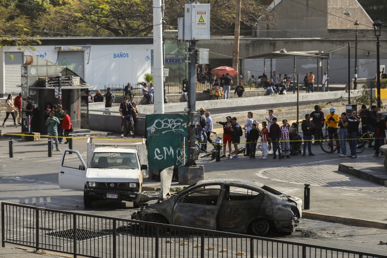 Two burned vehicles sit in the middle of a street as dozens of onlookers stand nearby behind yellow police tape.