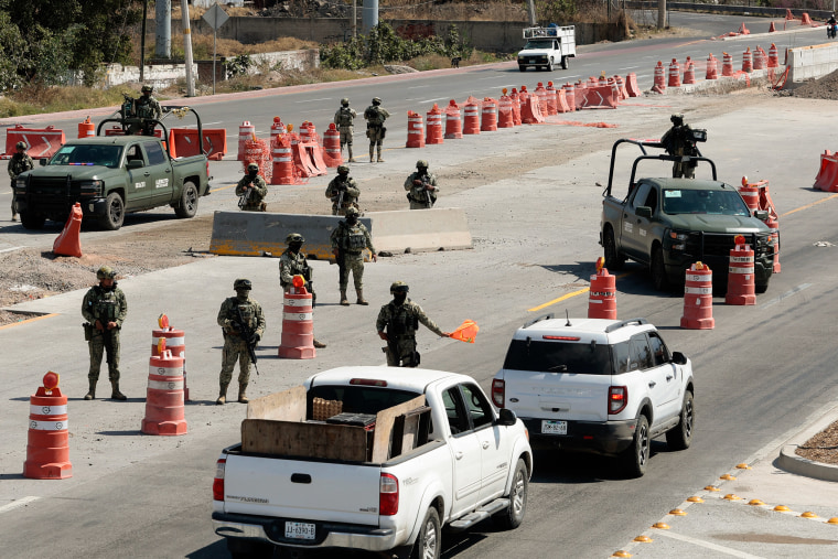 Soldiers wearing camouflage fatigues and holding rifles stand guard at a makeshift roadblock. One of them waves an orange flag toward passing vehicles.