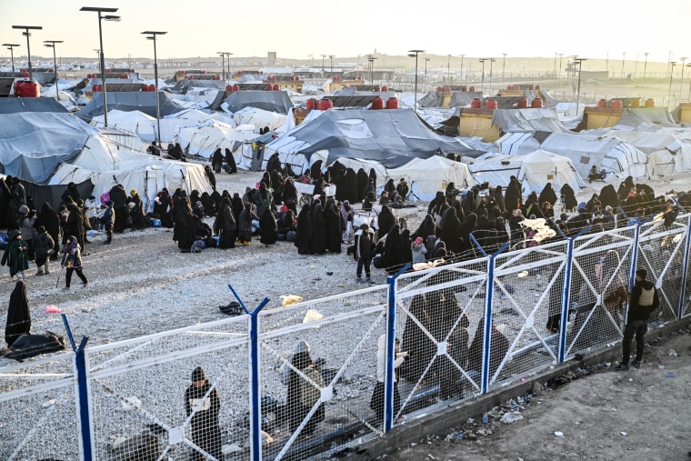 People stand around tents outside, a large guarded fence surrounds them