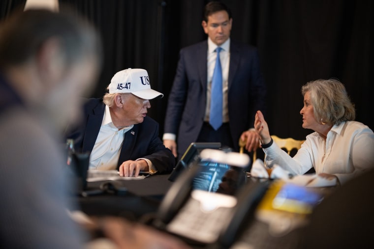Donald Trump sits at a conference table surrounded by presidential cabinet members. He is wearing a white USA cap.