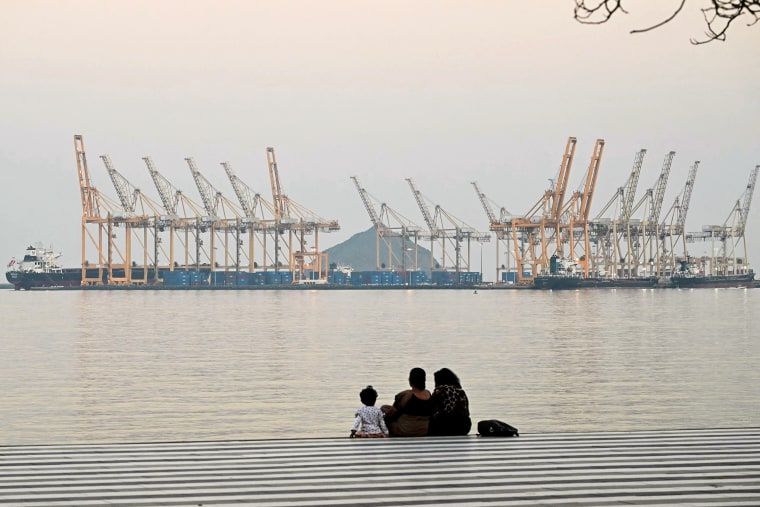 Two adults and one child sit on the ground across the water from dozens of maritime cranes and shipping containers. 