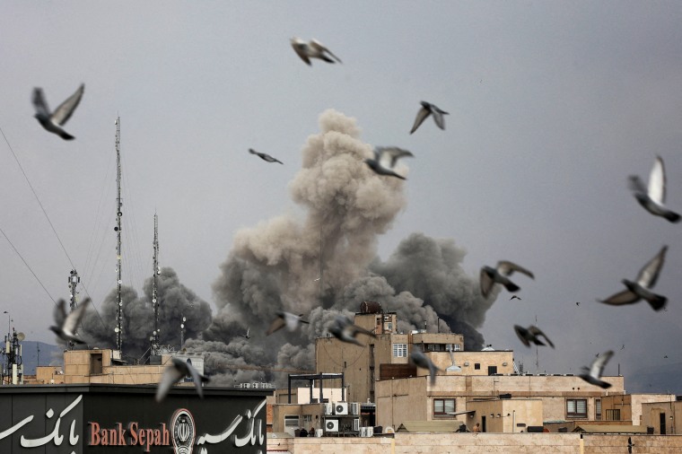 Birds fly as smoke rises between buildings.