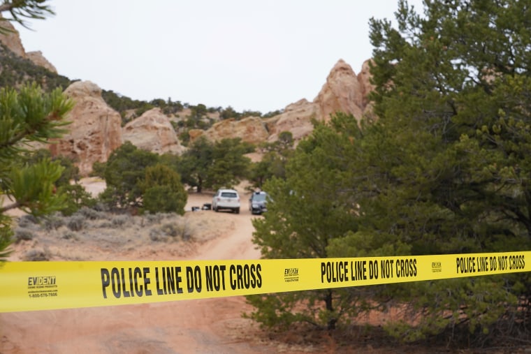 Yellow police tape stretches across a red dirt road leading toward two parked vehicles, surrounded by small jagged rock formations.