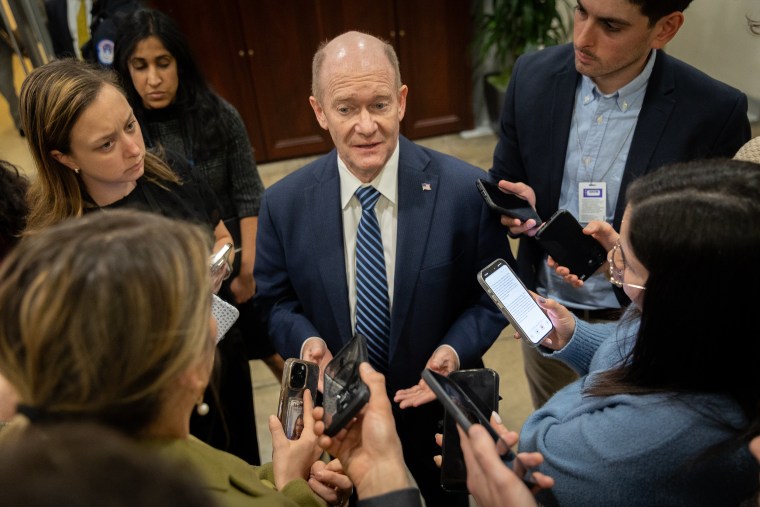 Chris Coons stands surrounded by reporters holding their phones out toward him.
