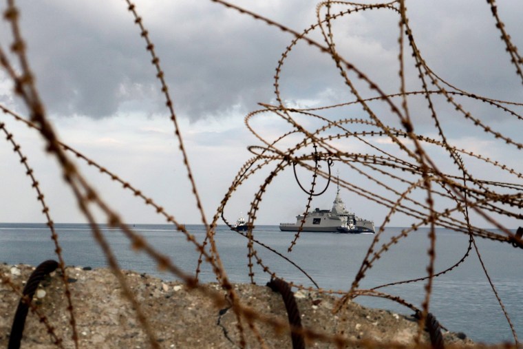 A military ship is visible through serpentine wire tangled along a rocky shoreline.