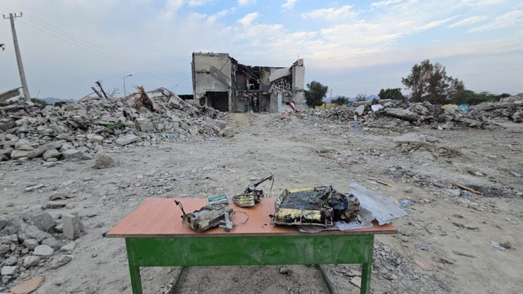 Missile debris is gathered on a table outside, a destroyed building is seen is the distance