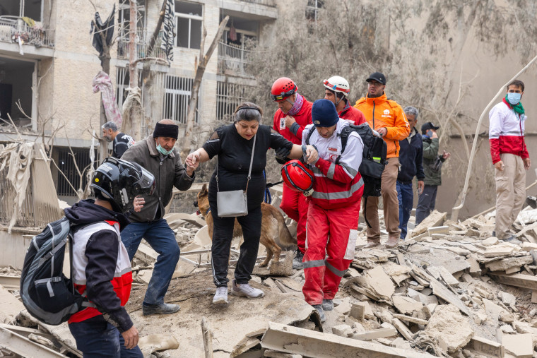A woman is helped through debris by rescue workers following a strike on a residential building on March 16, 2026 in central Tehran, Iran.