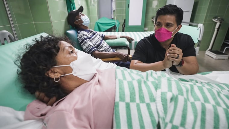 George Solis, right, wearing a pink mask, speaks with Yonelkys García who is laying in a hospital bed and wearing a white mask