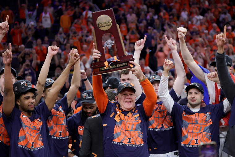O time de basquete da Universidade de Illinois apoia seu treinador enquanto ele segura um troféu acima de sua cabeça.