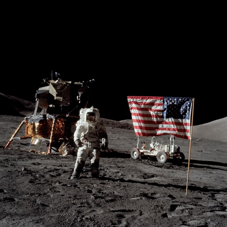 Harrison Schmitt está na superfície da Lua em um traje espacial, com uma bandeira americana afixada no chão à sua direita e um piloto do módulo lunar à sua esquerda.