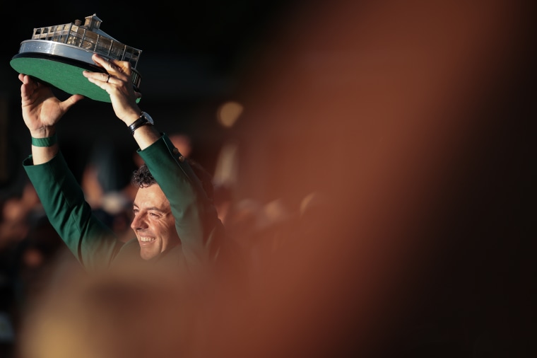 Rory McIlroy, wearing the champion's green jacket, holds up a trophy and smiles.