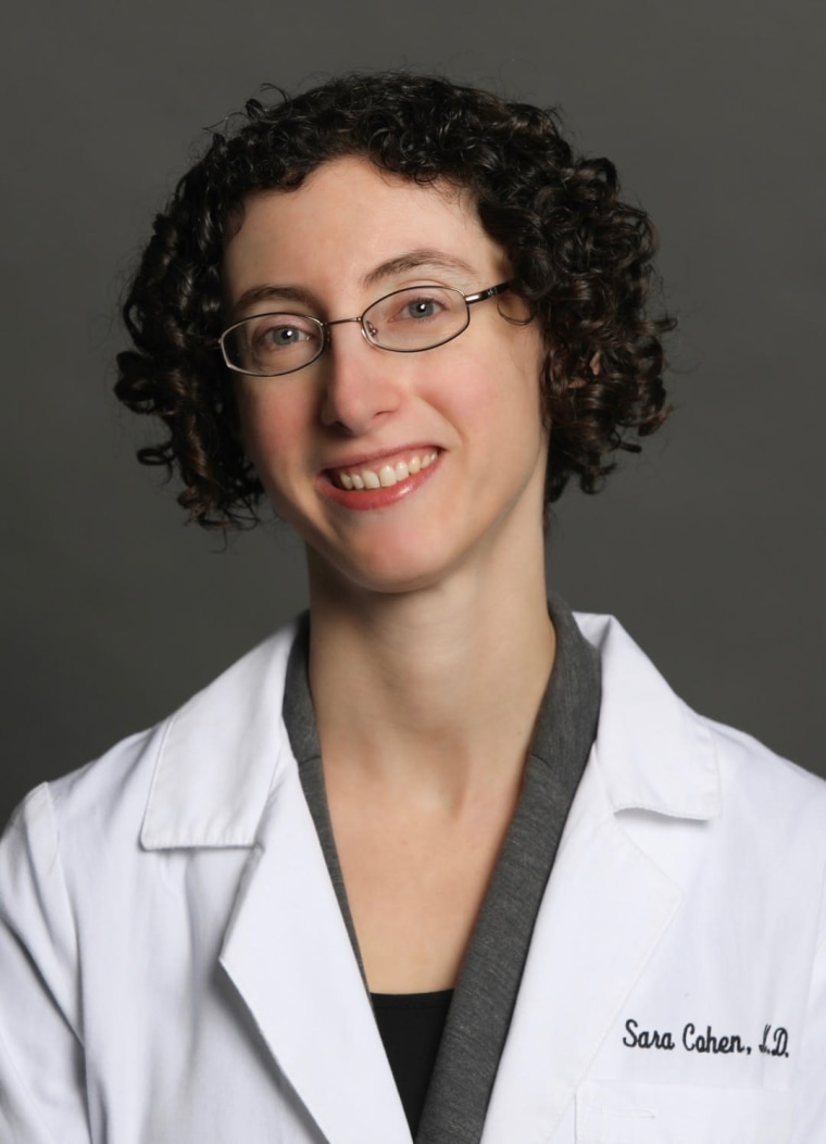 Η Freida McFadden χαμογελά για ένα πορτρέτο σε σκούρο γκρι φόντο, φορώντας ένα λευκό παλτό εργαστηρίου με μαύρο ραμμένο κείμενο που γράφει "Sara Cohen"