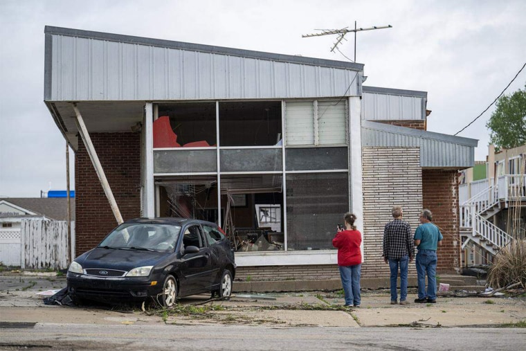 People stand outside of a damaged building with a damaged car parked out front