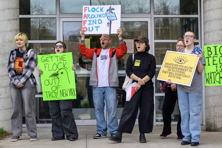 Six people hold signs in front of a glass doorway during a protest against surveillance cameras.