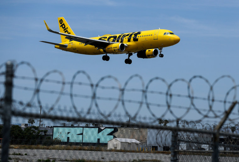 A yellow Spirit Airlines planes can be seen flying above a barbed wire fence in the foreground