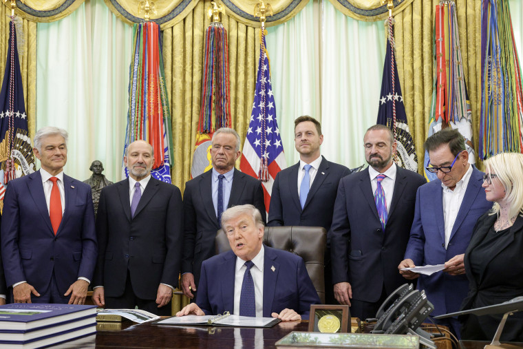 Donald Trump seated at his desk in the Oval Office surrounded by cabinet members. 