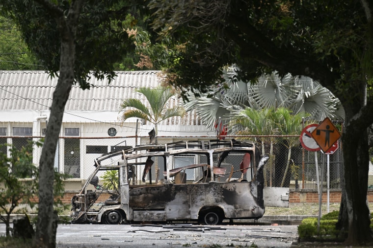 A charred, burned-out bus in front of a one-story building with metal roof, surrounded by palmetto trees.