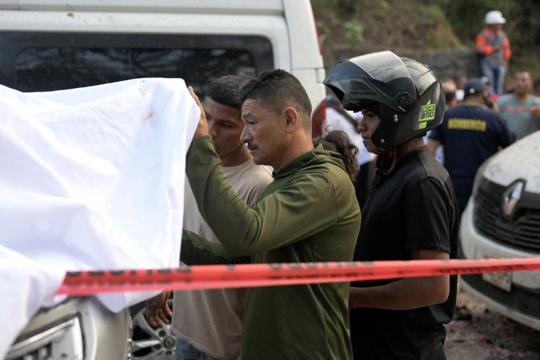 A man lifts a white sheet covering a body as two other men look on, behind red crime scene tape.