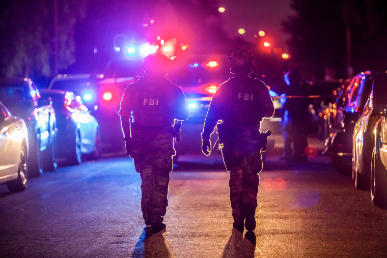 Two FBI field agents wearing helmets and camouflage fatigues walk in front of a vehicle with red and blue flashing lights.