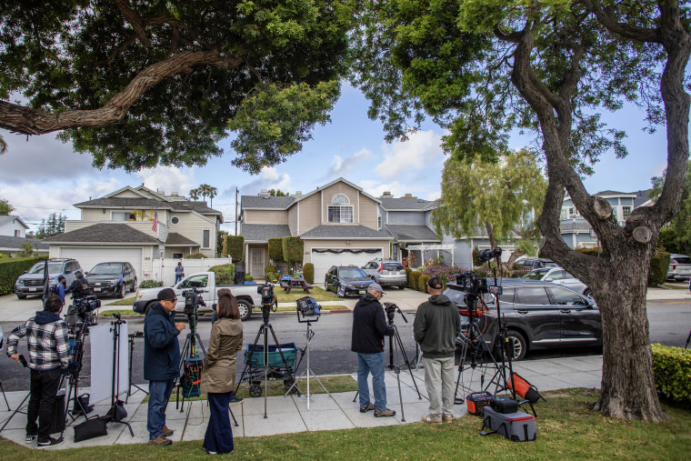 A line of reporters with television cameras stand across the street from a row of two story residential homes on a tree-lined street.