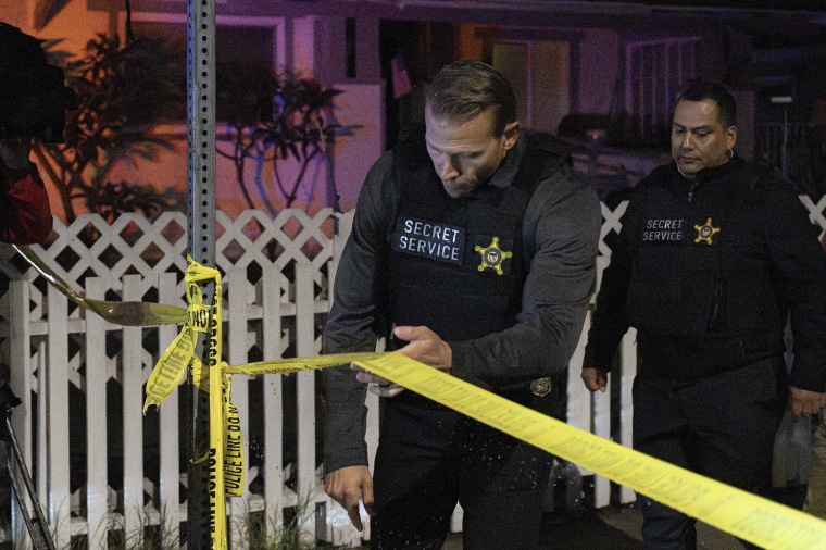 Two field agents wearing black flak jackets with Secret Service patches on them cross a yellow police perimeter.