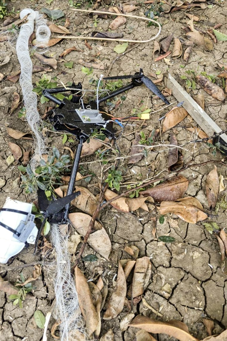 A broken drone surrounded by a thick tangle of wires lying on a patch of dry, cracked earth.