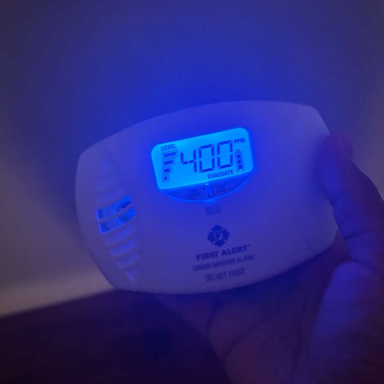 Person holding First Alert CO615 Dual-Power Plug-In Carbon Monoxide Detector with right hand in the dark and the blue backlit screen reading “Evacuate” and “400.”