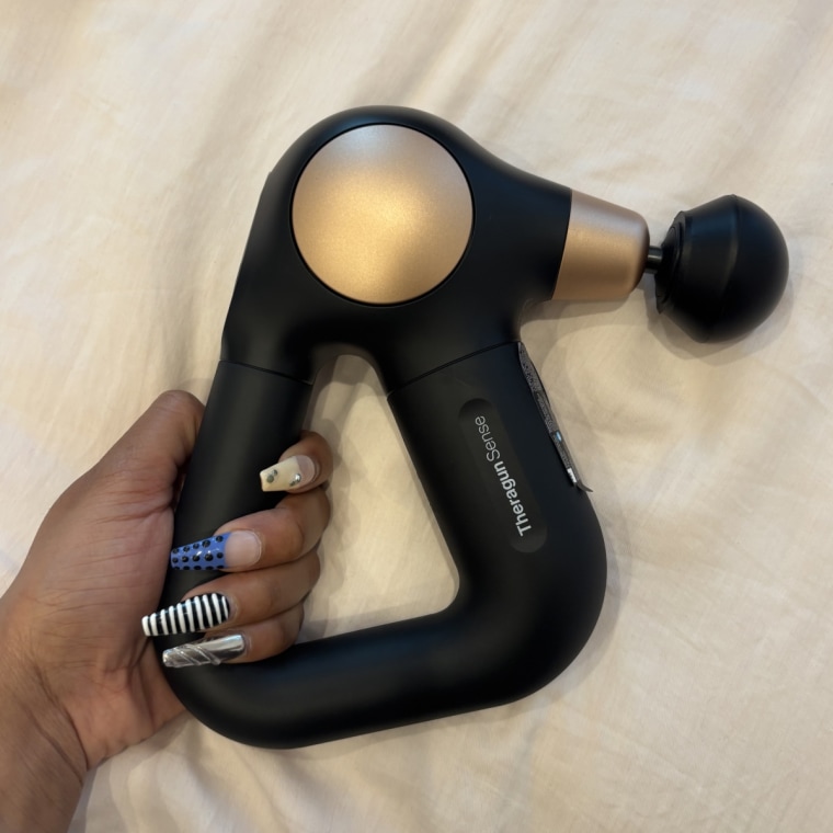 A hand holds a Therabody massage gun in front of a white background.