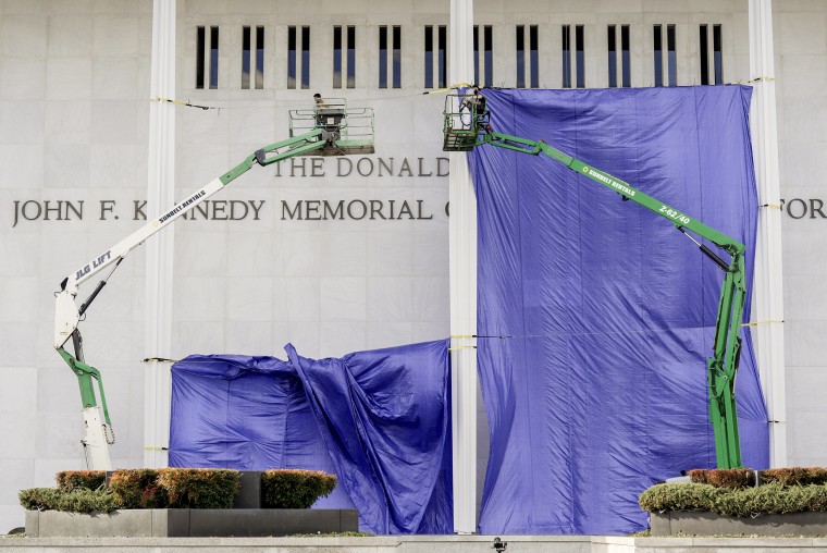 Workers in two hydraulic cranes are suspended above the ground in front of the Kennedy Center's facade, with a giant purple tarp stretched behind them.