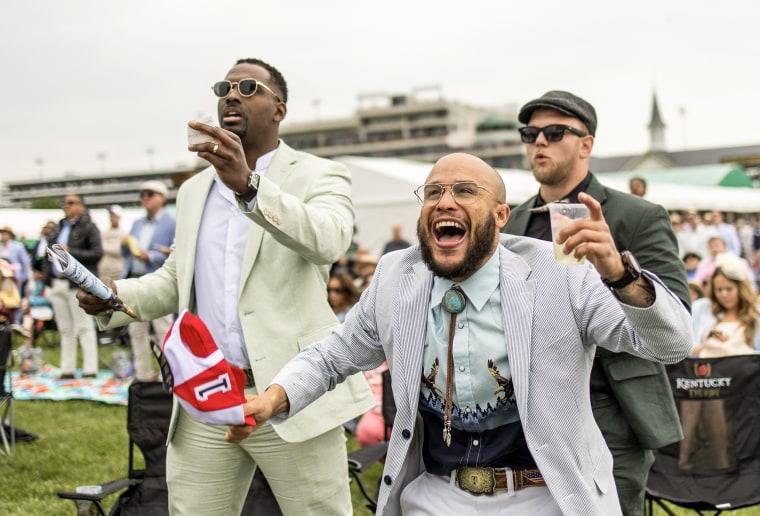 Three men cheer while standing in the infield of Churchill Downs under gray skies.