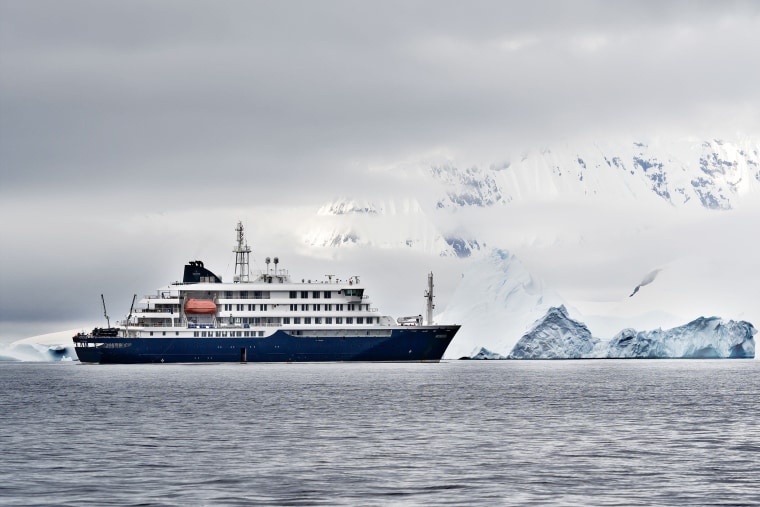 A cruiser ship in arctic waters with a large iceberg shrouded in clouds behind it