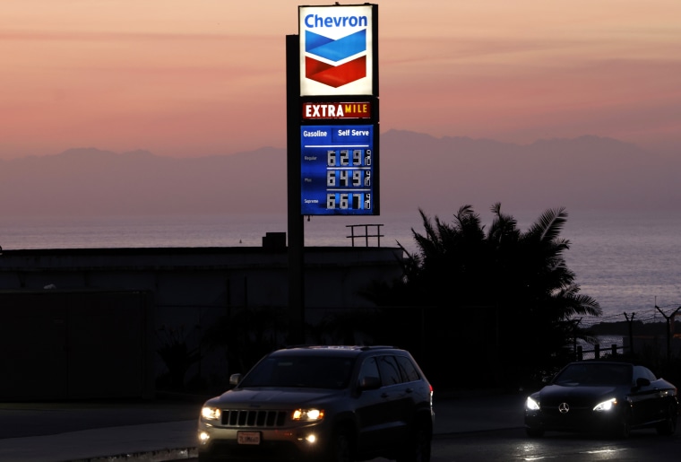 A Chevron gas station sign against a sunset sky with cars driving past.