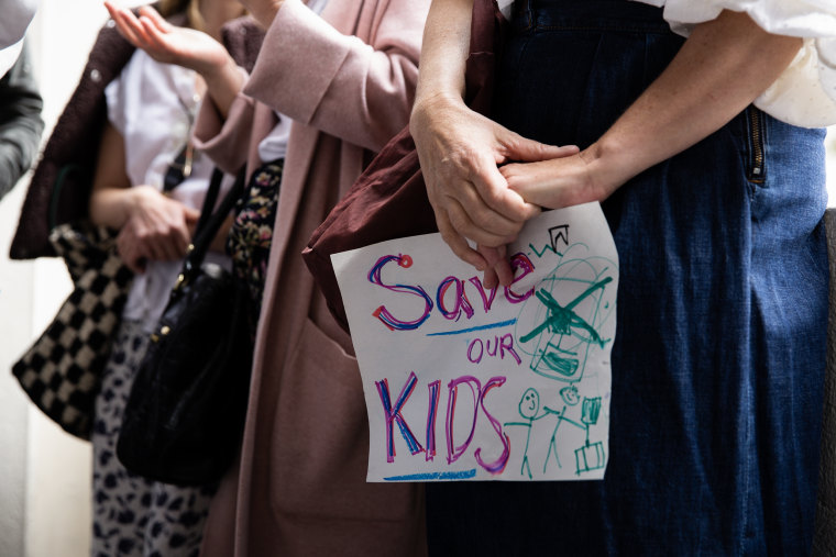 A person seen from the waist down holding a hand-written sign reading Save Our Kids. 
