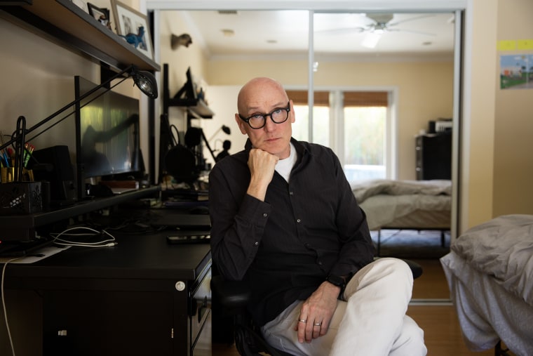 A bald man with black eyeglasses rests his chin on one hand while seated at a desk in a bedroom.