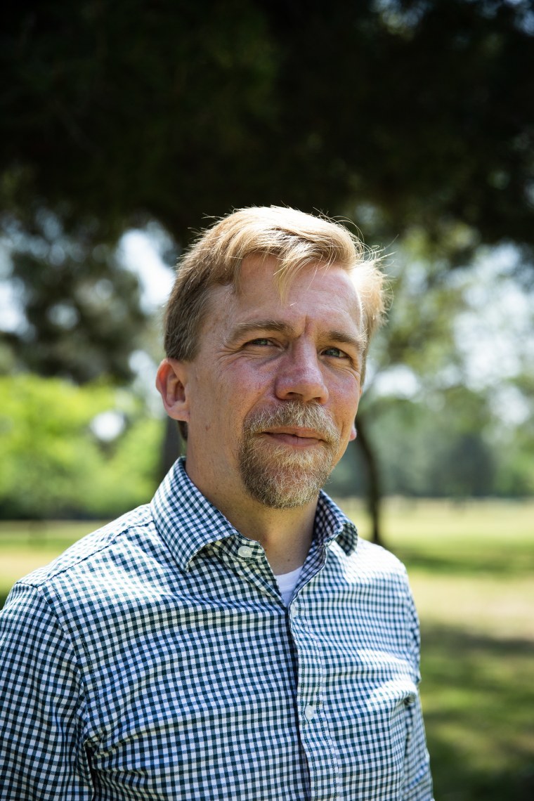 A goateed man wearing a checkered shirt standing for a portrait in the shade.