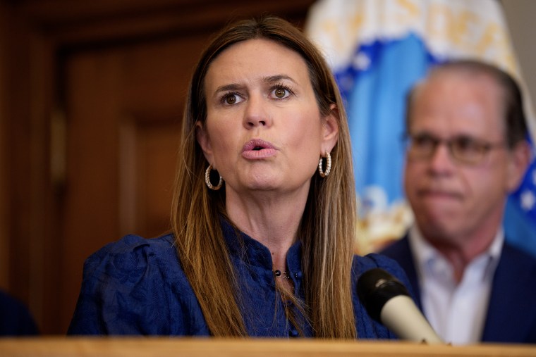 Sarah Huckabee speaking at a lectern with a man standing in soft focus behind her.