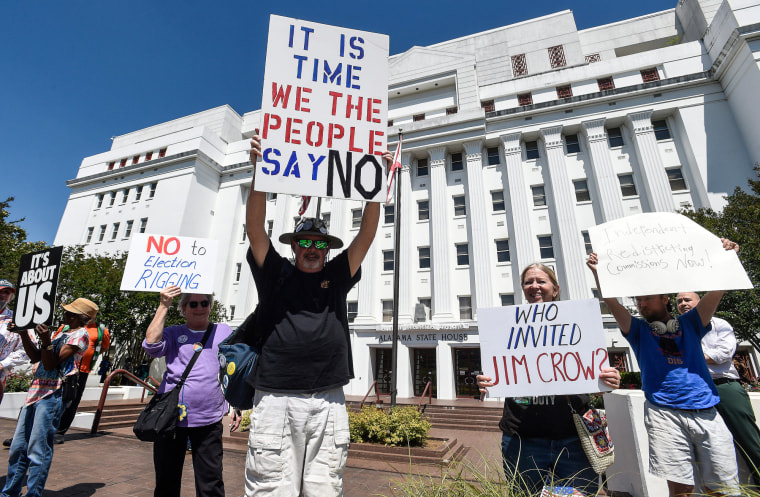 People hold signs outside against redistricting, slogans such as "It's About Us" / "No to Election Rigging" / "It is time we the people say no" / and "Who invited Jim Crow?" can be seen written on signage