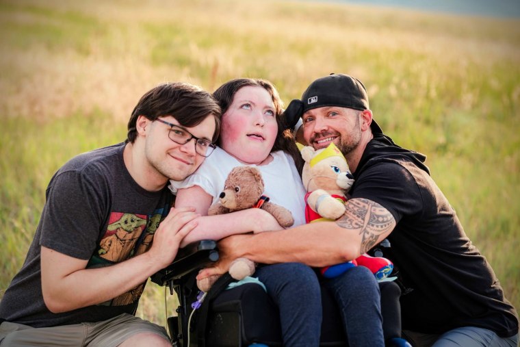 Casey Barrett and his son hug his daughter, center, who is seated in a wheelchair, she holds two teddy bears, they are all in a field