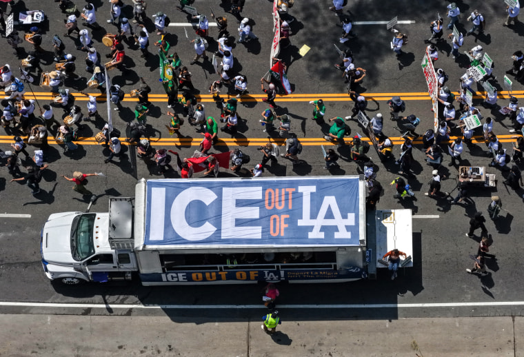 A aerial view of a truck with a banner on top that reads "ICE out of LA", driving next to a large group protestors walking down the street